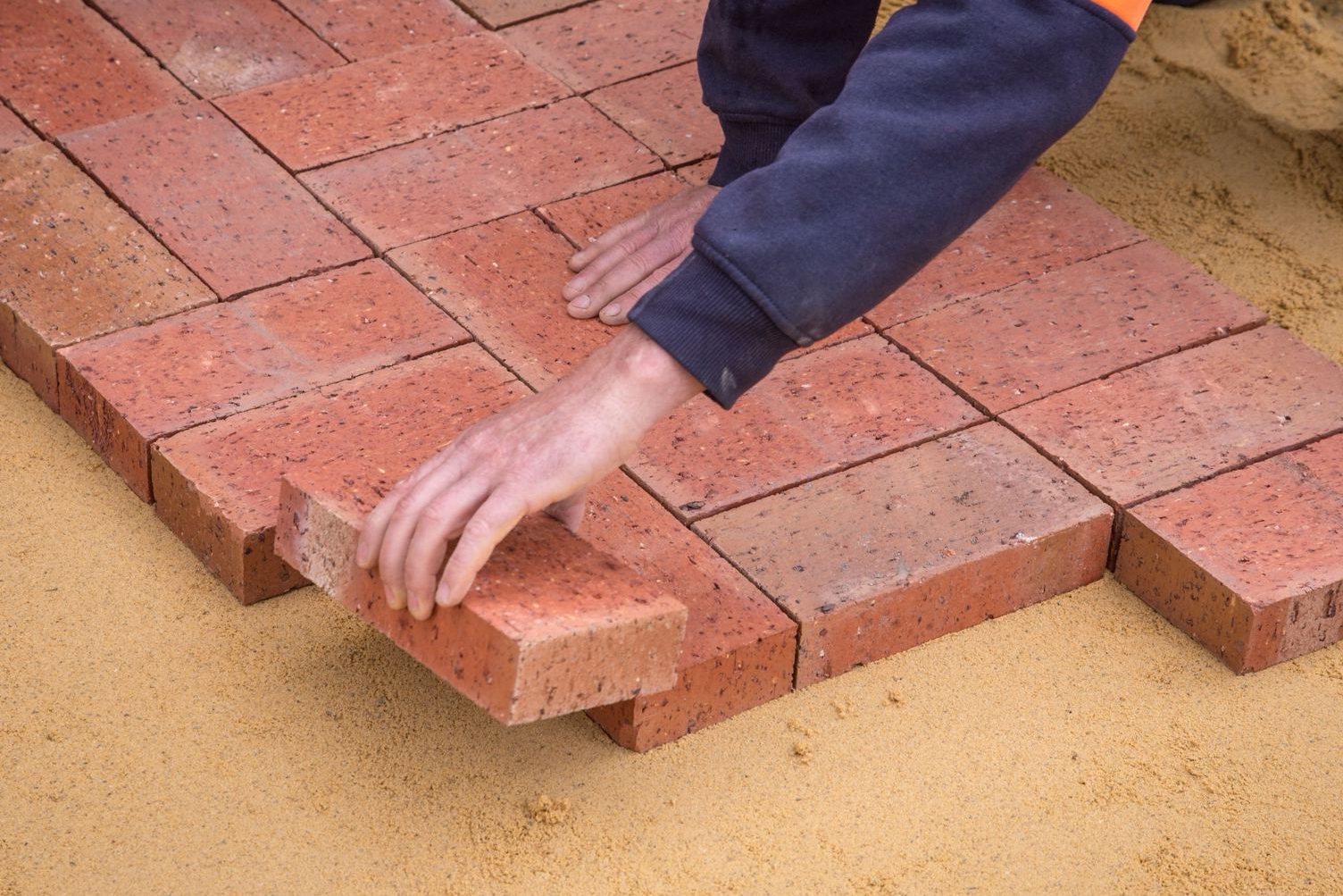Close-up of hands laying red brick pavers on a bedding sand base