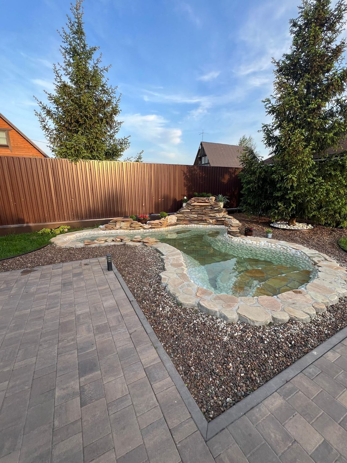 Stone-edged backyard pond beside a paver patio with tall pine trees behind